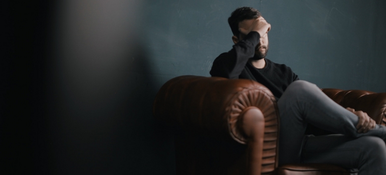 A depressed man with a hand on a head sitting on the sofa and thinking about fitness for anxiety and depression.