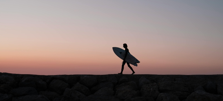 Person walking with surfboard at sunset along a rocky breakwater.